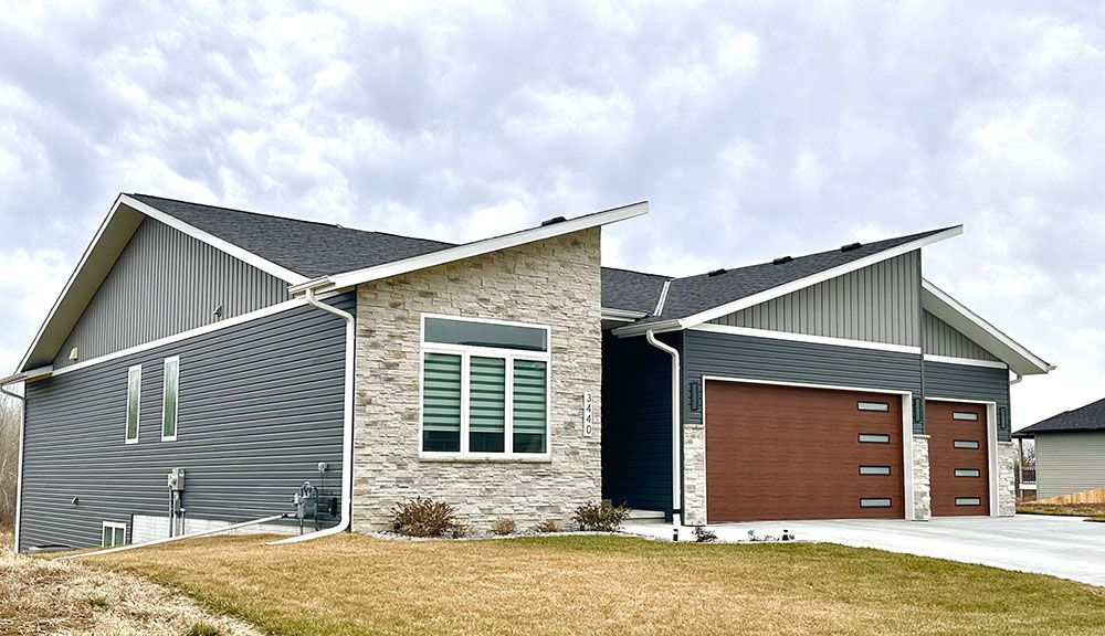 Modern one-story house with gray and brick facade, brown garage doors, and a cloudy sky.