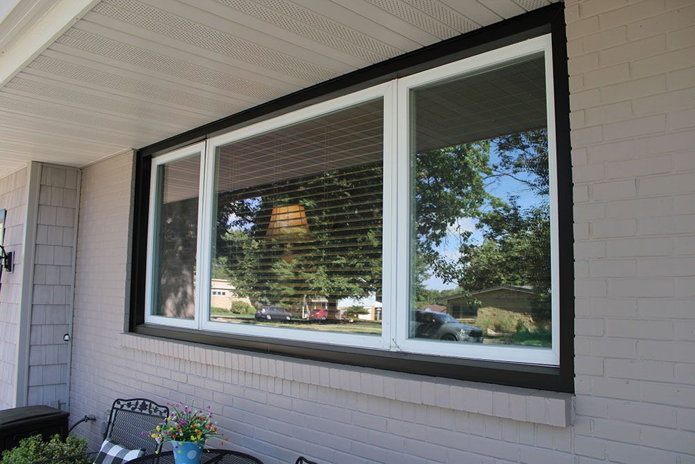 Large window with black trim, reflecting trees and sky, on a gray brick exterior.