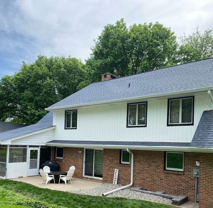 Two-story white house with brick base, blue roof, black-framed windows, and a patio with furniture under a cloudy sky.