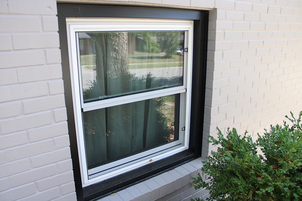 White-framed window in a gray brick wall, partially open. Black trim surrounds the window. A bush is in the foreground.
