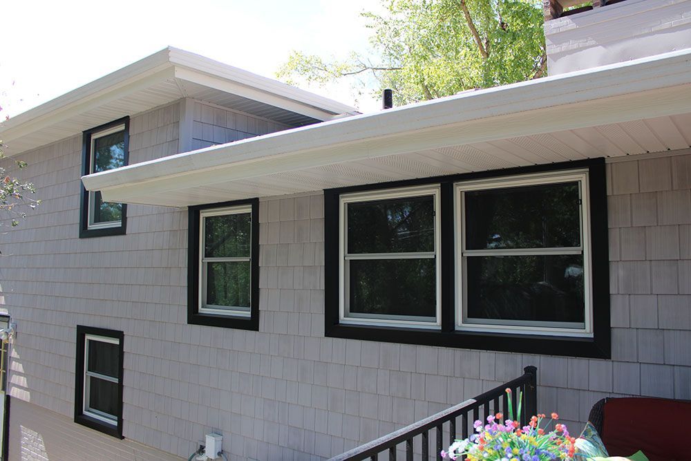 Gray shingle house with black-framed windows and white trim. Overhanging roof and small railing.