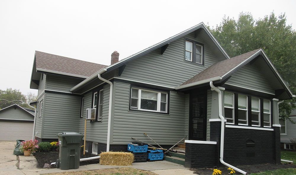 A two-story house with green siding, brown roof, and porch. Trash cans and a hay bale are in the yard.