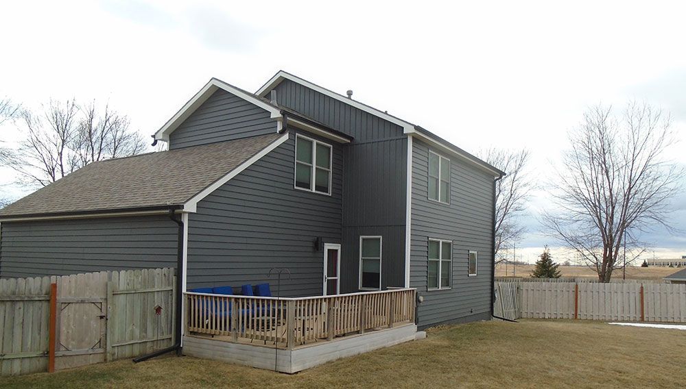 Back of a two-story gray house with a wooden deck and a brown fence in a yard with sparse grass.