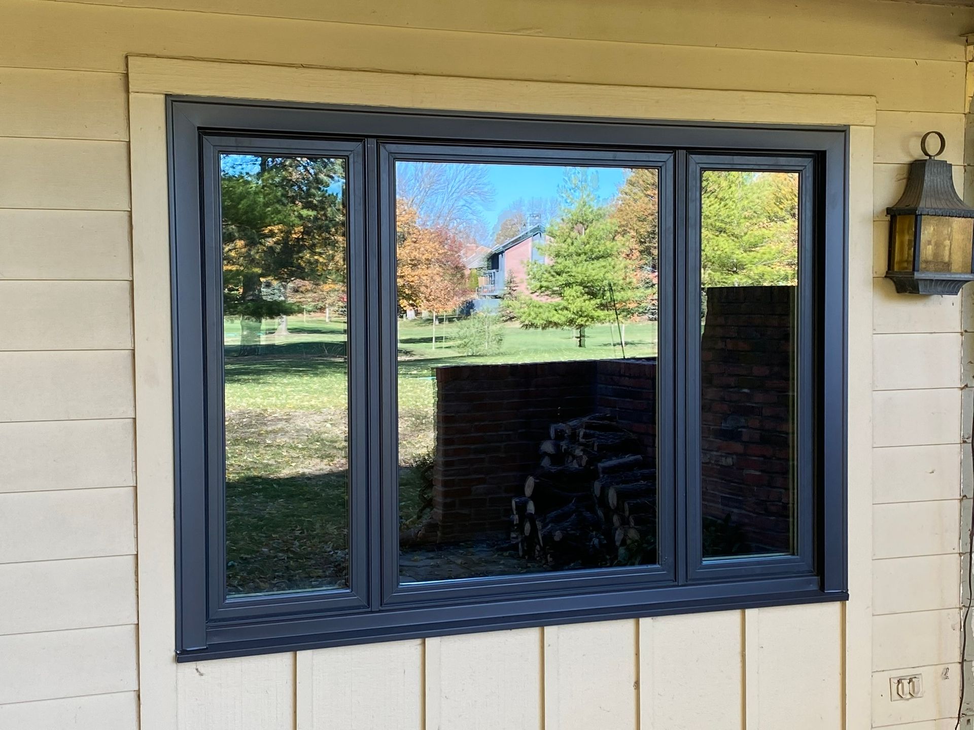 Dark-framed window with reflections of trees and house, set in a light-colored exterior wall.