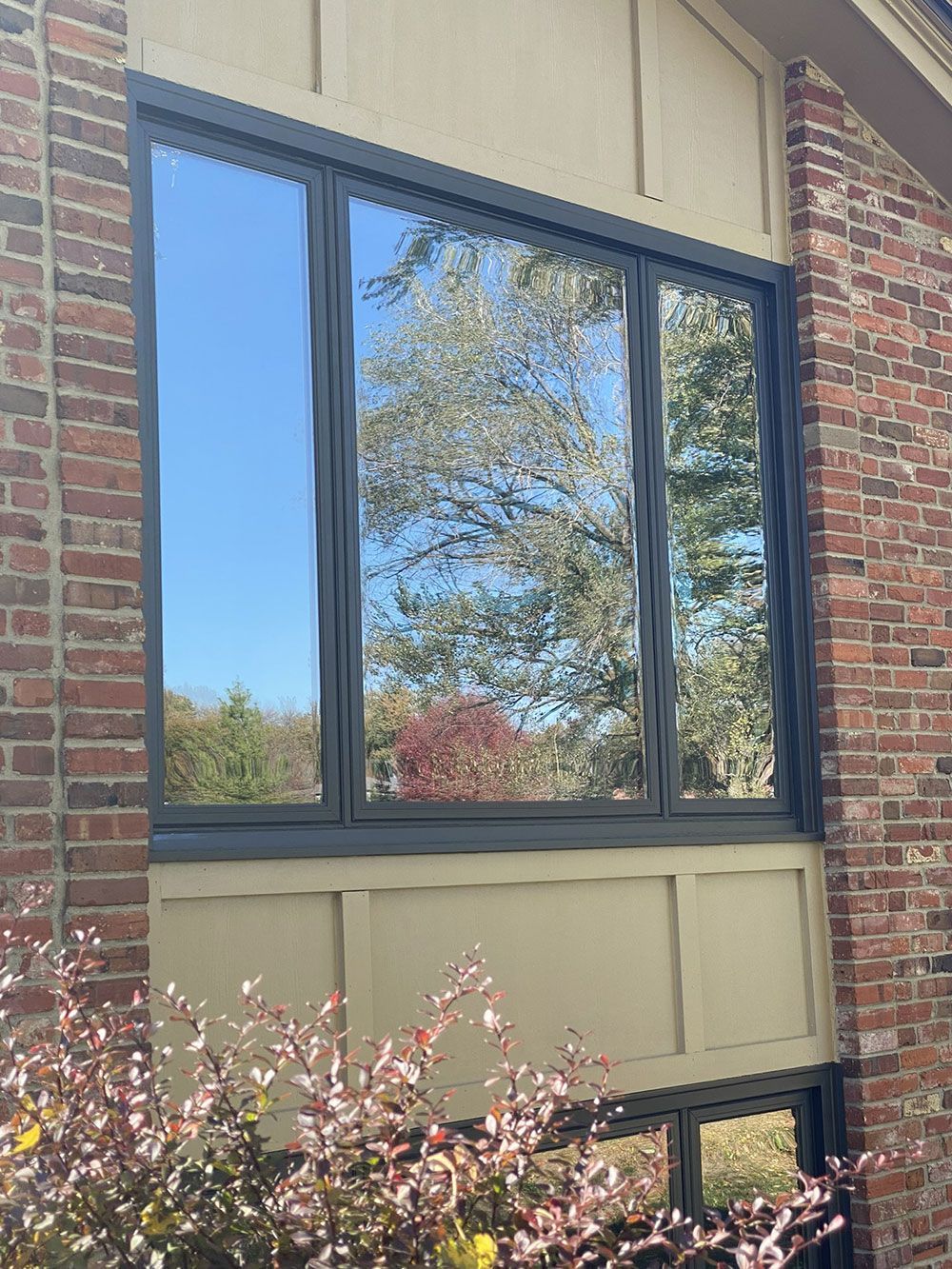 A multi-pane window reflects a clear blue sky and trees, framed by a dark trim on a brick building.