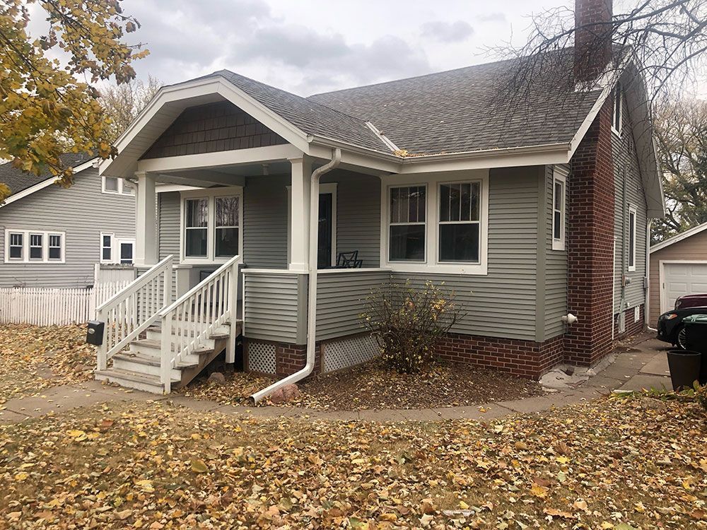 A one-story house with gray siding, white trim, and a brick chimney under a cloudy sky.