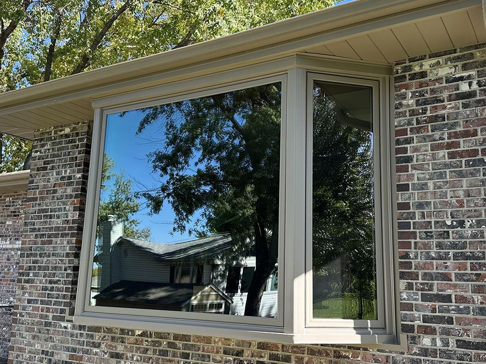 Bay window in brick wall, reflecting trees and a house, beige trim.