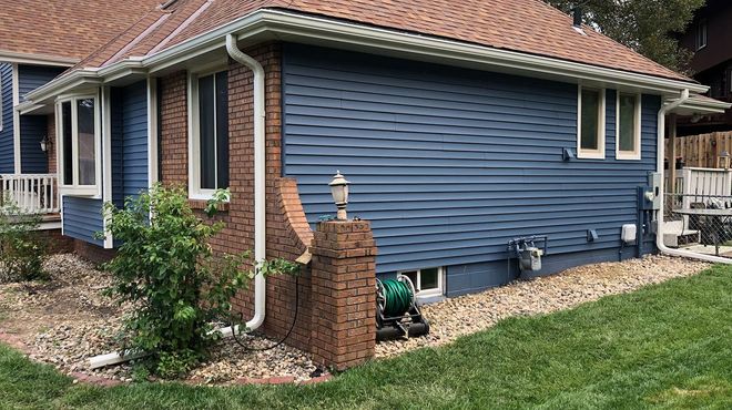 House exterior with blue siding, brick chimney, and white gutters. Green lawn and gravel landscaping.