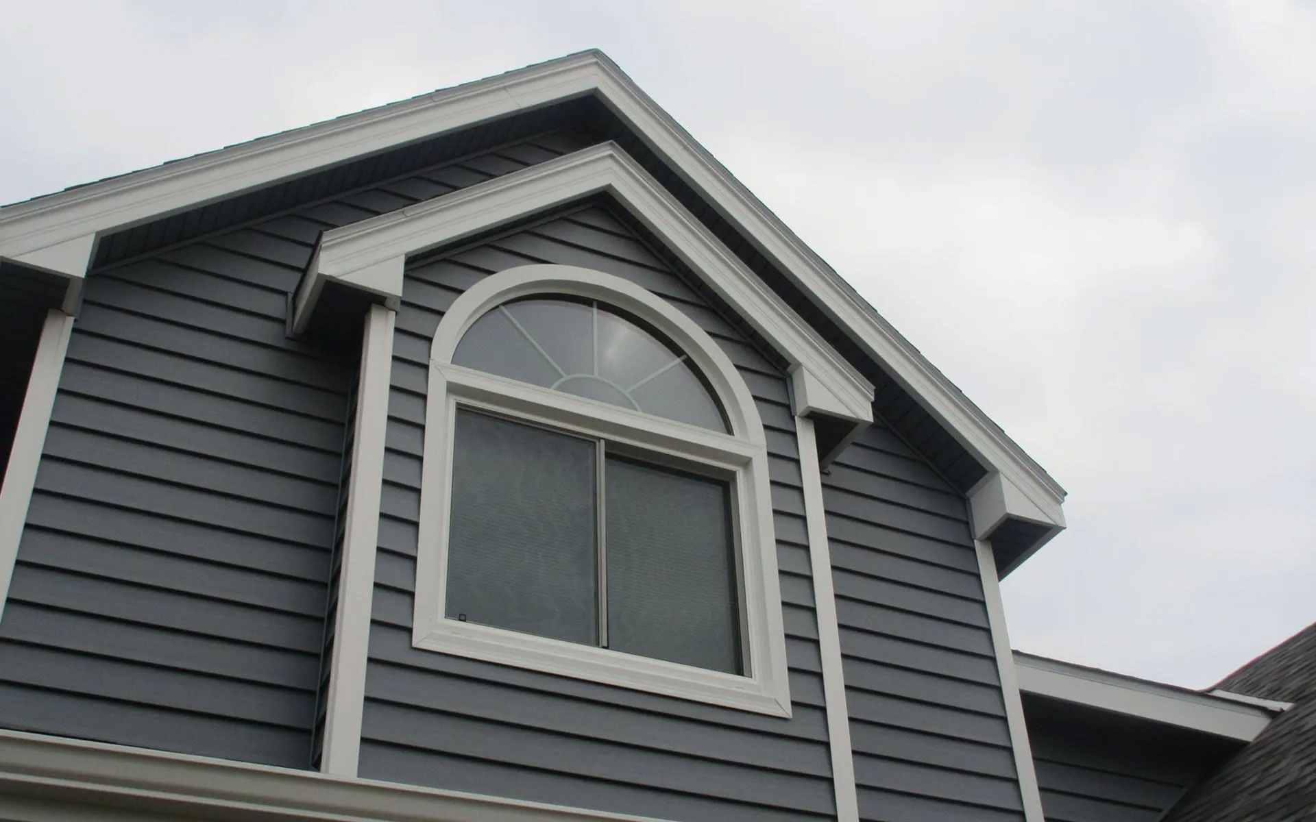 Blue-sided house with a white-framed arched window under a triangular peak, against a cloudy sky.