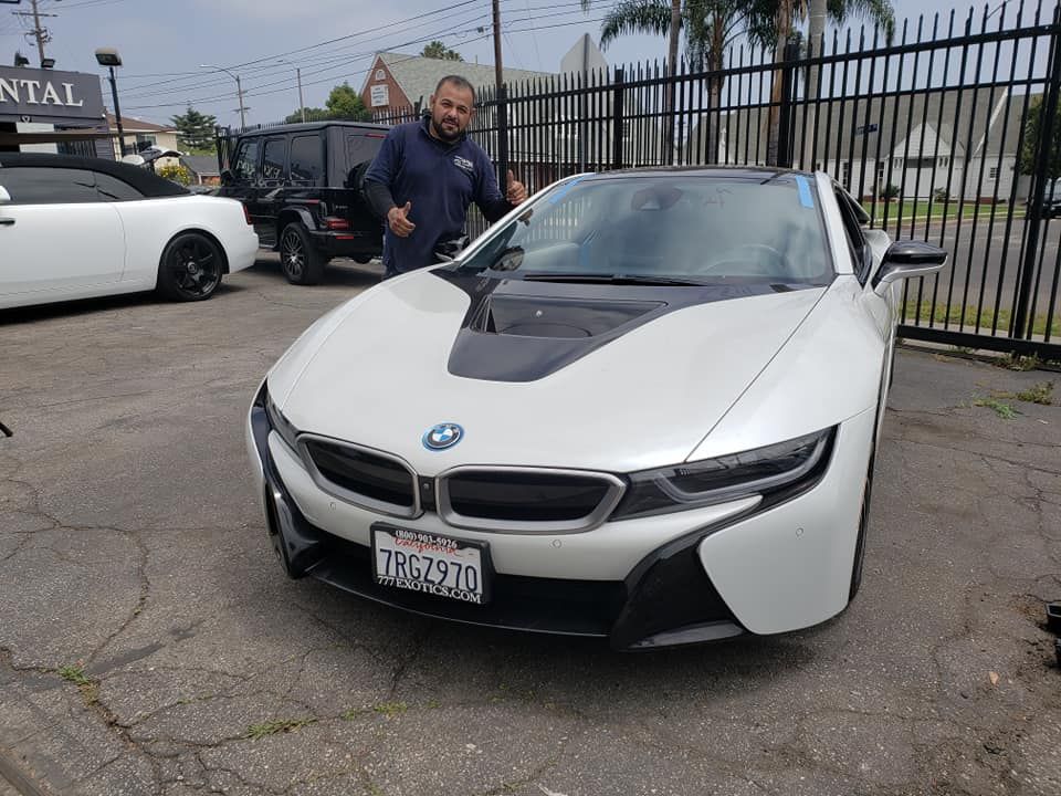 Man gives thumbs up next to a white BMW i8. Car is parked in lot.
