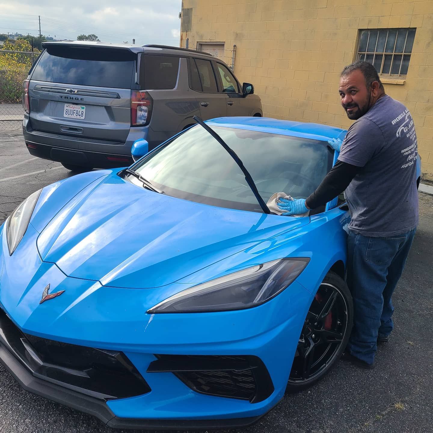 Man cleaning the windshield of a bright blue sports car. Grey SUV and yellow building in the background.