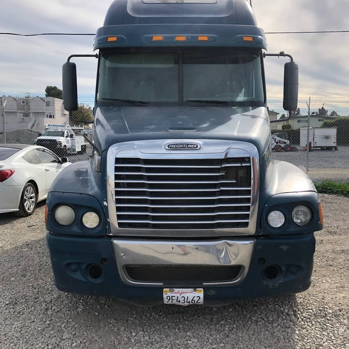 Blue Freightliner semi-truck parked outdoors. Gray bumper, chrome grill, and California license plate visible.