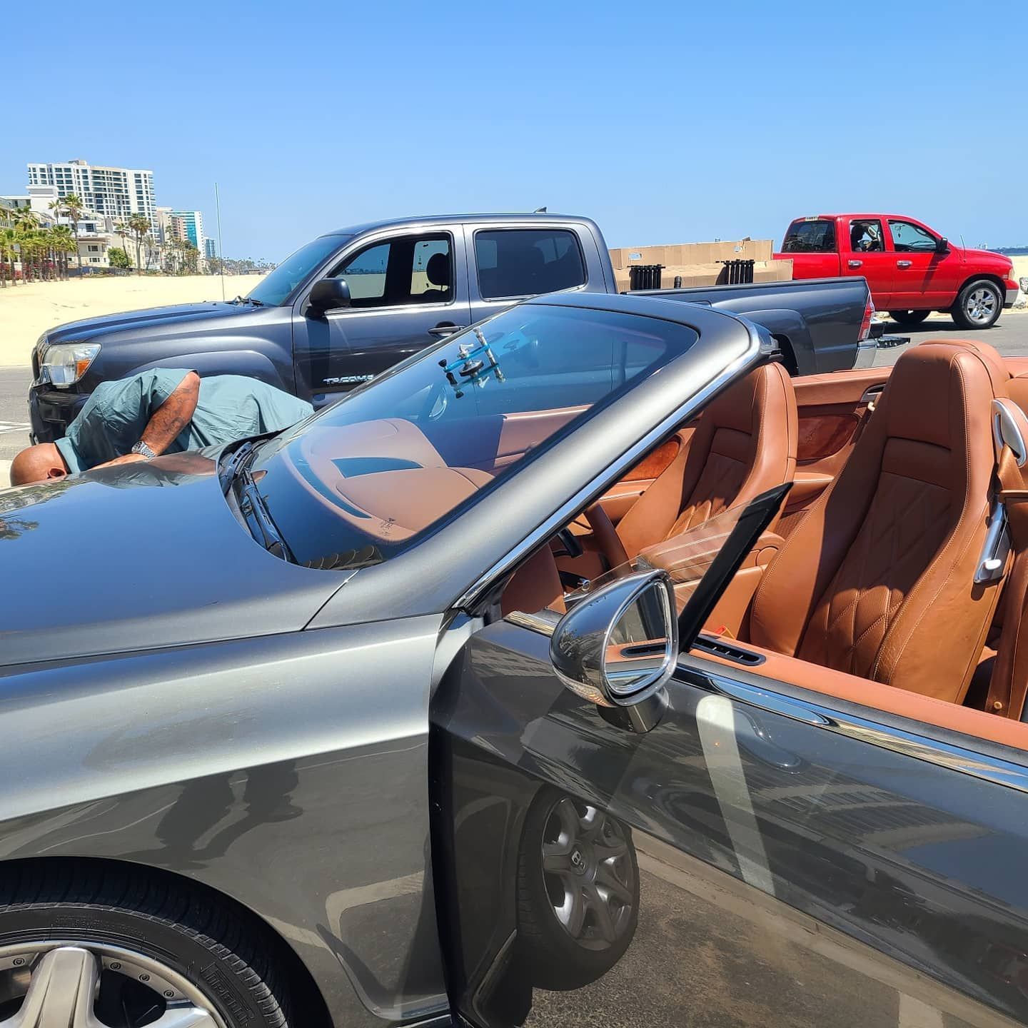 Grey convertible car with tan interior; two people nearby, two trucks in background, beach setting.
