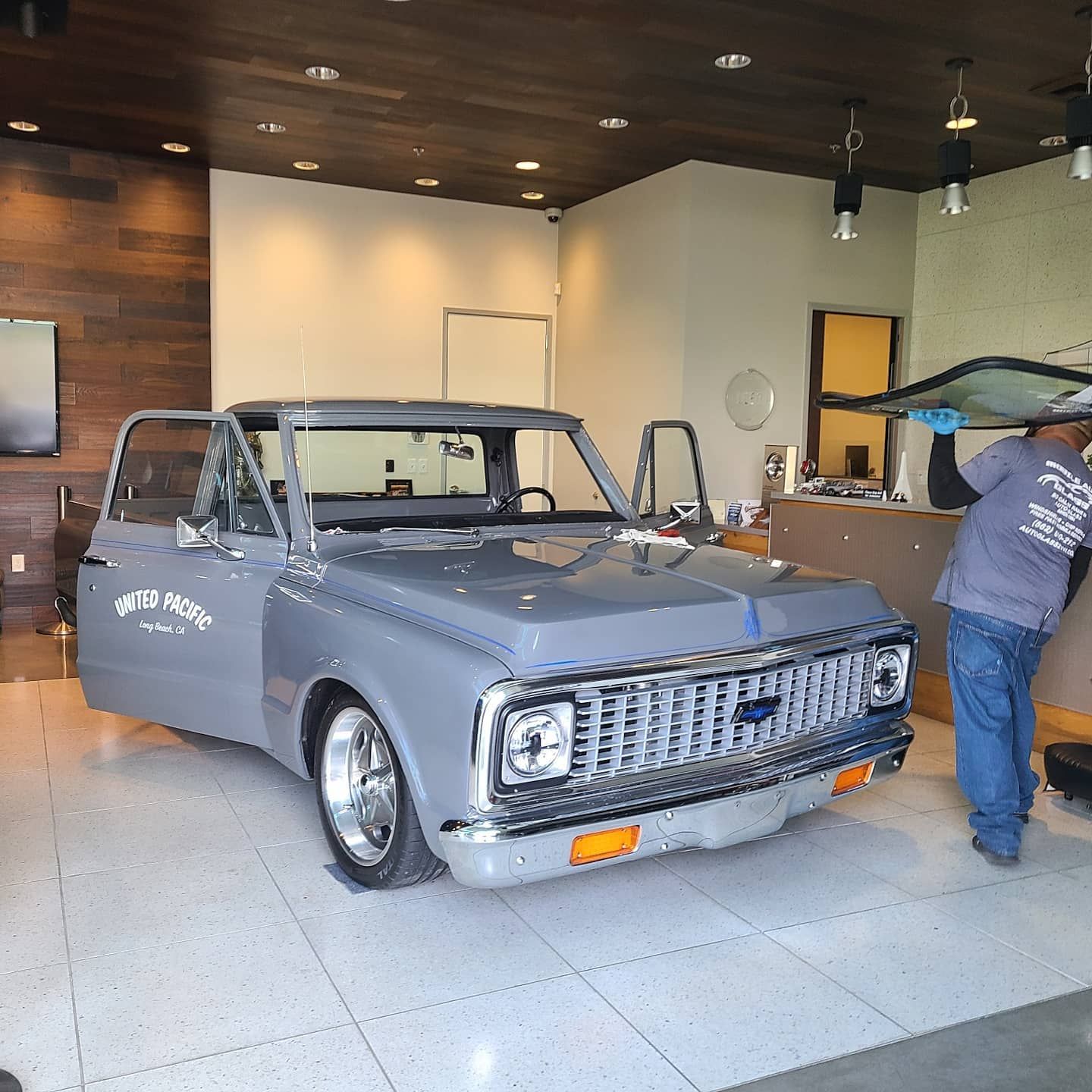 A gray classic Chevrolet truck with open doors, being worked on inside a shop. A worker is handling a window.