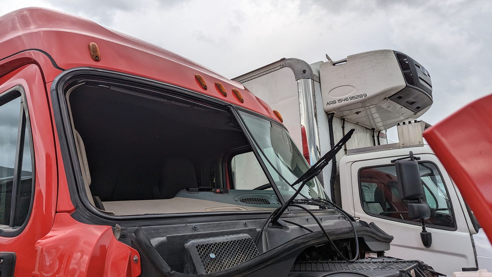 Red semi-truck cab with broken windshield, white refrigerated trailer in background. Cloudy sky.