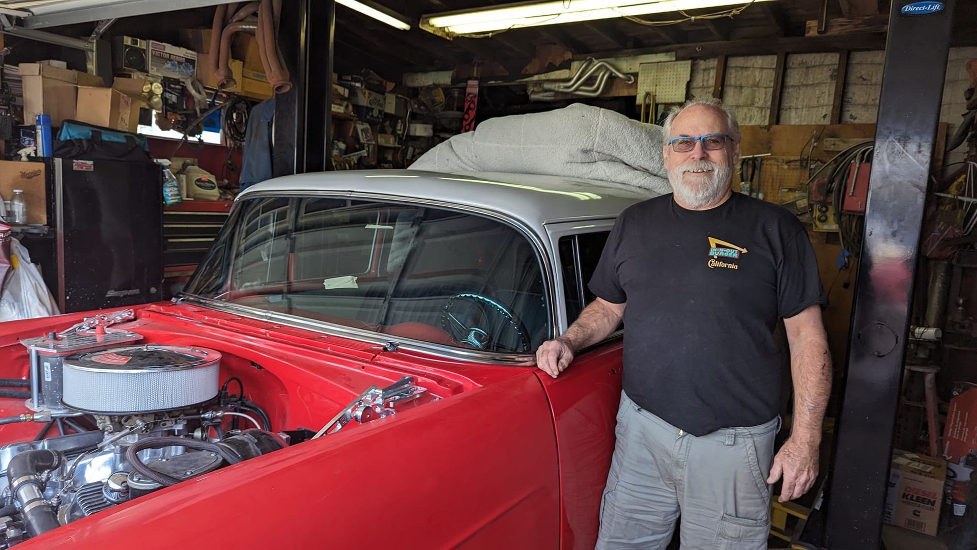 Man standing by a red classic car in a garage; engine visible.