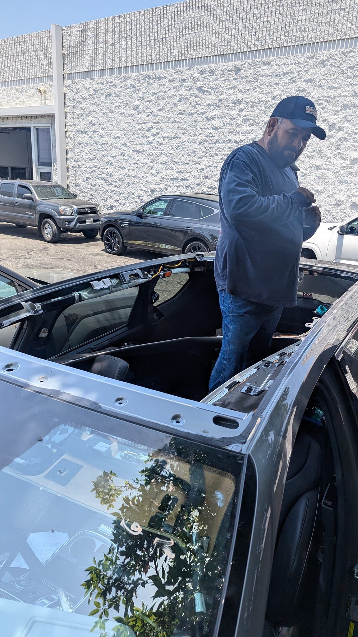 Man standing inside car, looking down. Black car with open roof. Cars and building in background.