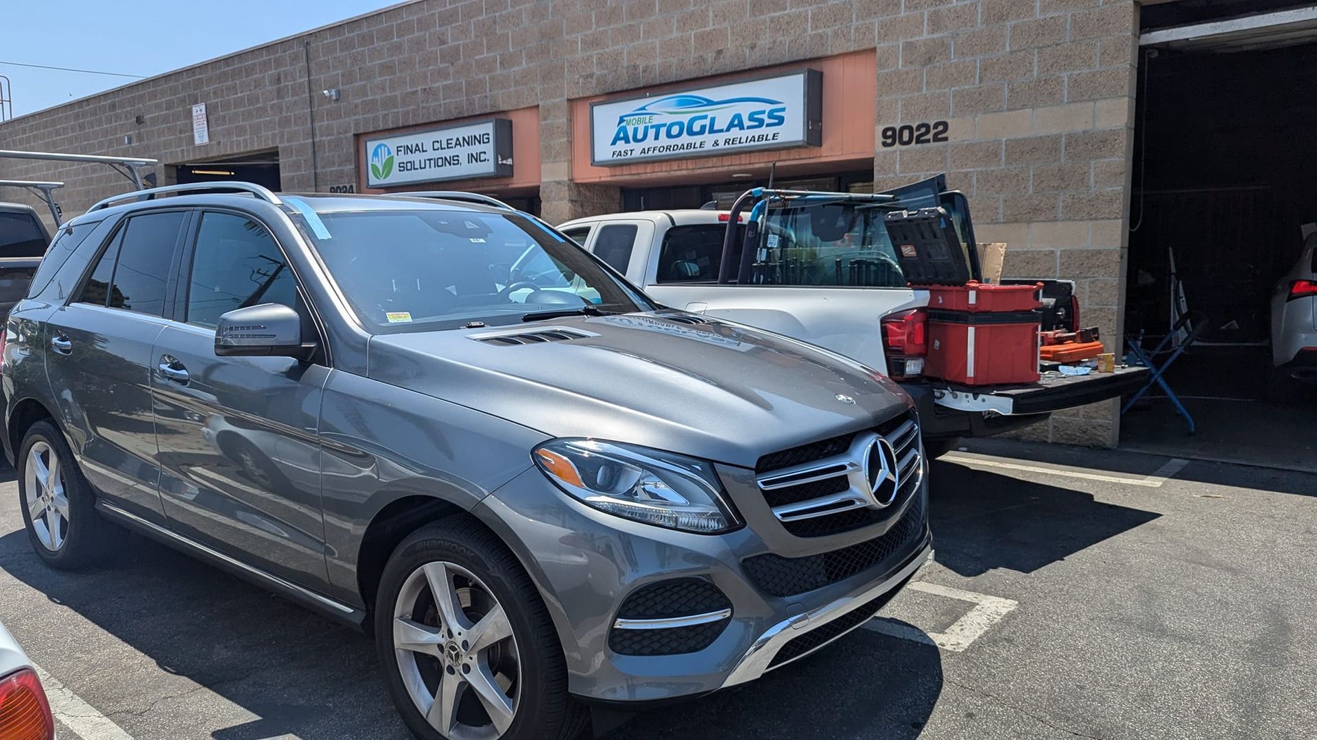Gray Mercedes SUV parked in front of an auto glass repair shop; another vehicle with an open bed is behind it.