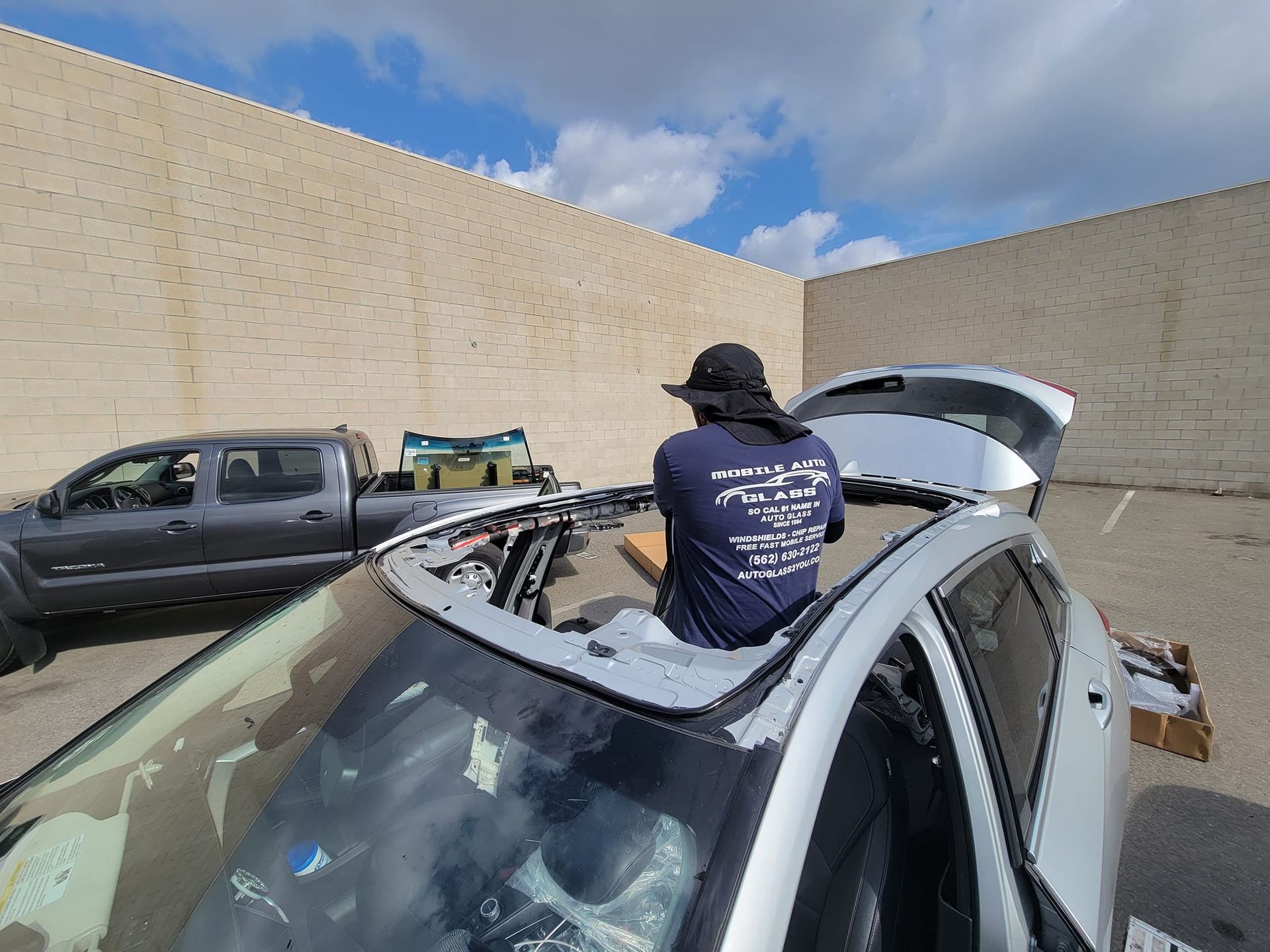 Person working on a car sunroof in a parking lot. Silver car, gray truck, brick wall background.