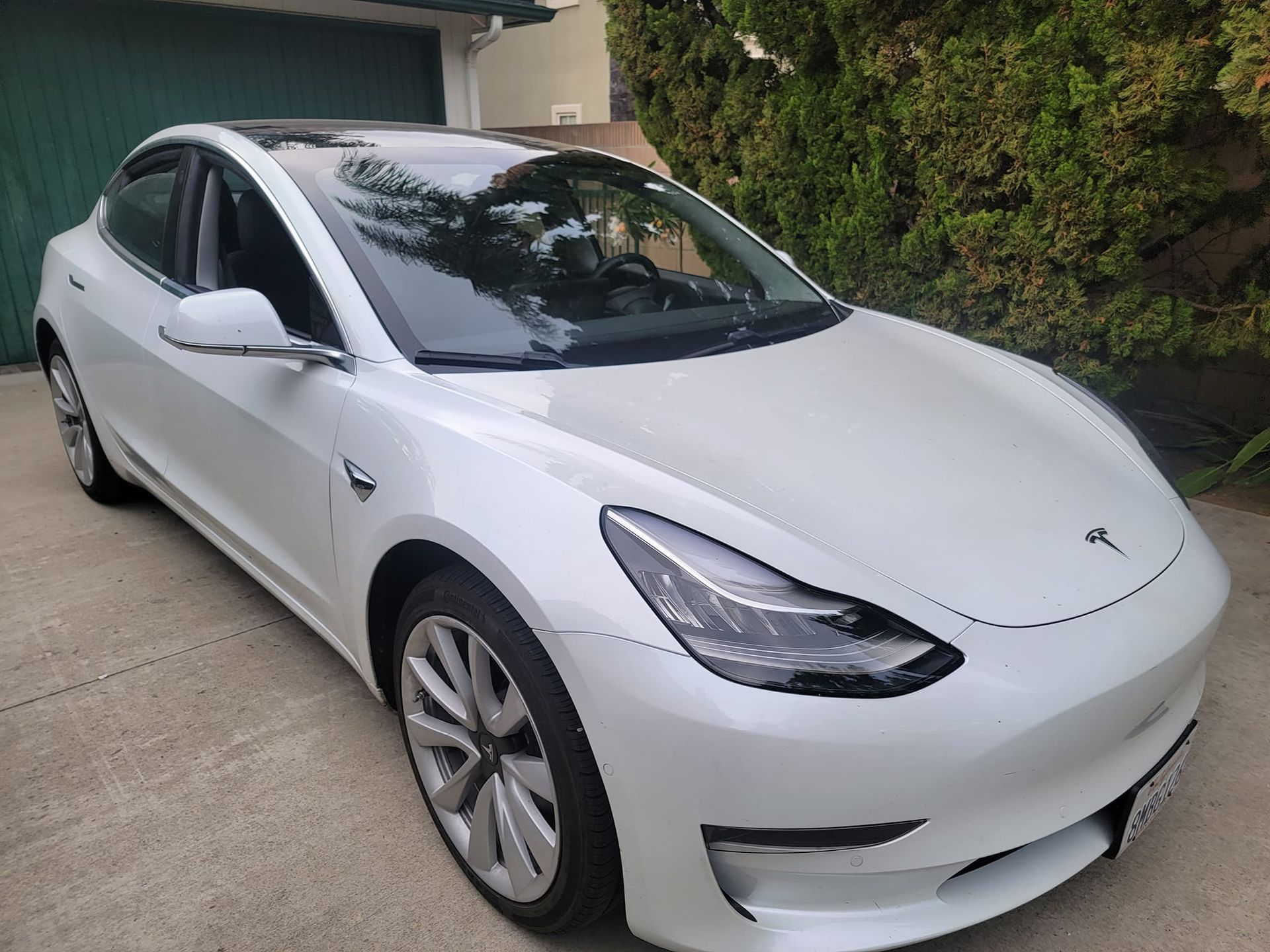 White Tesla Model 3 car parked on a paved driveway with greenery in the background.