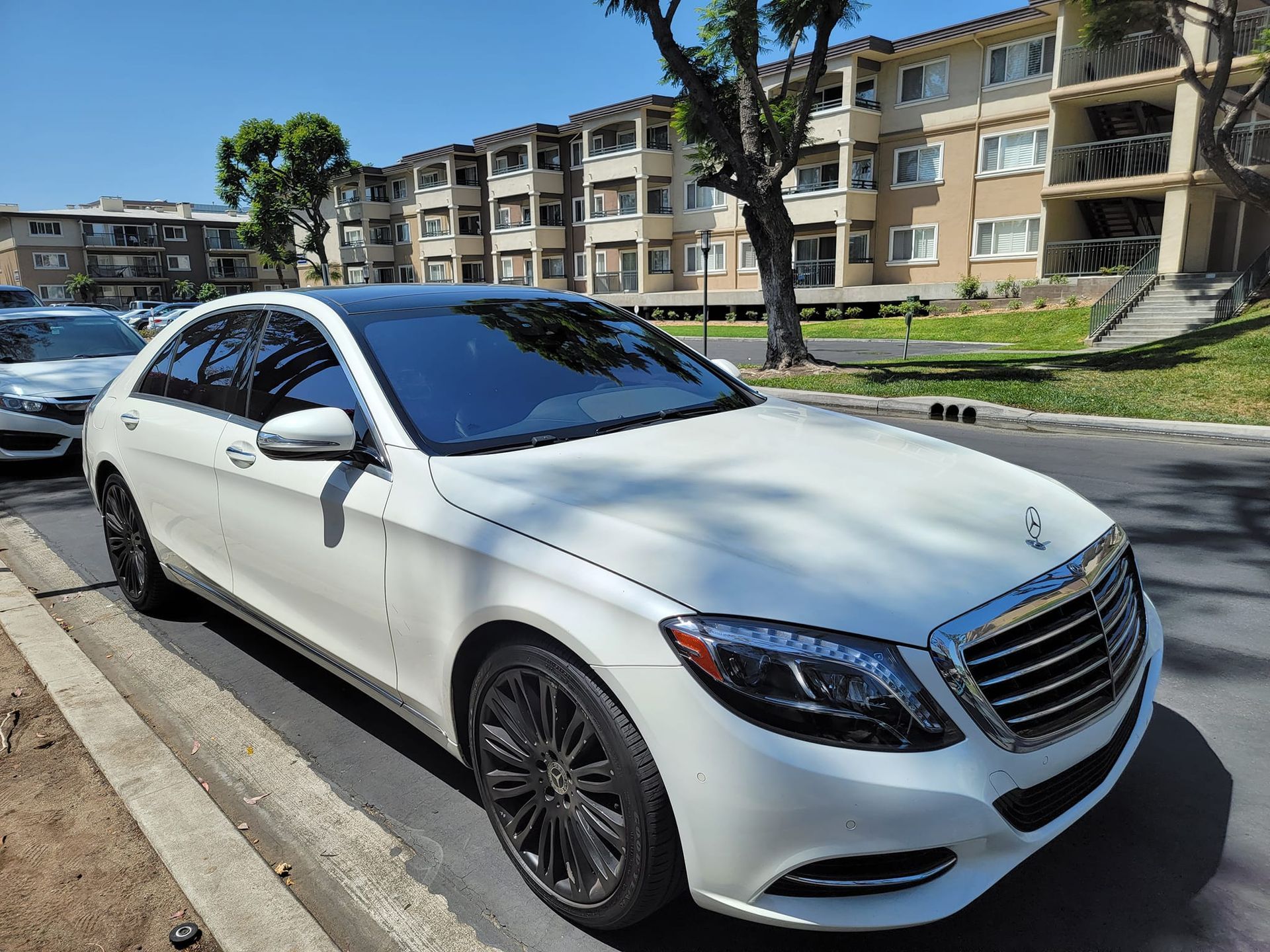 White Mercedes-Benz sedan parked on a street in front of a multi-story apartment building on a sunny day.