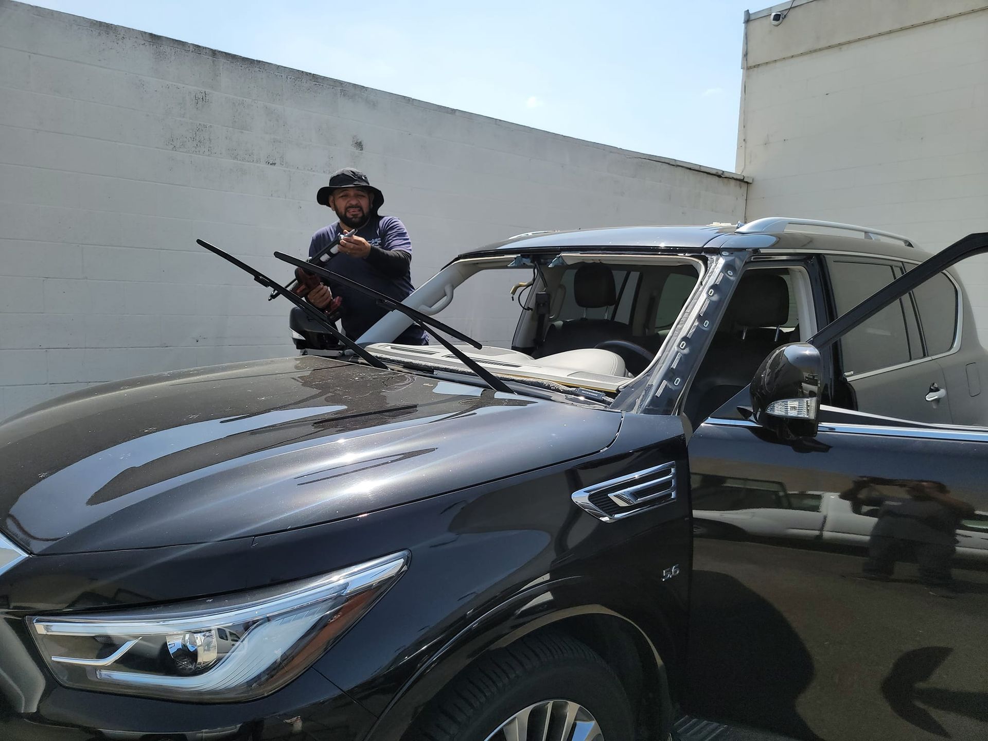 Man standing by damaged SUV windshield, holding what appears to be a gun. Car's door is open. Bright, outdoor setting.