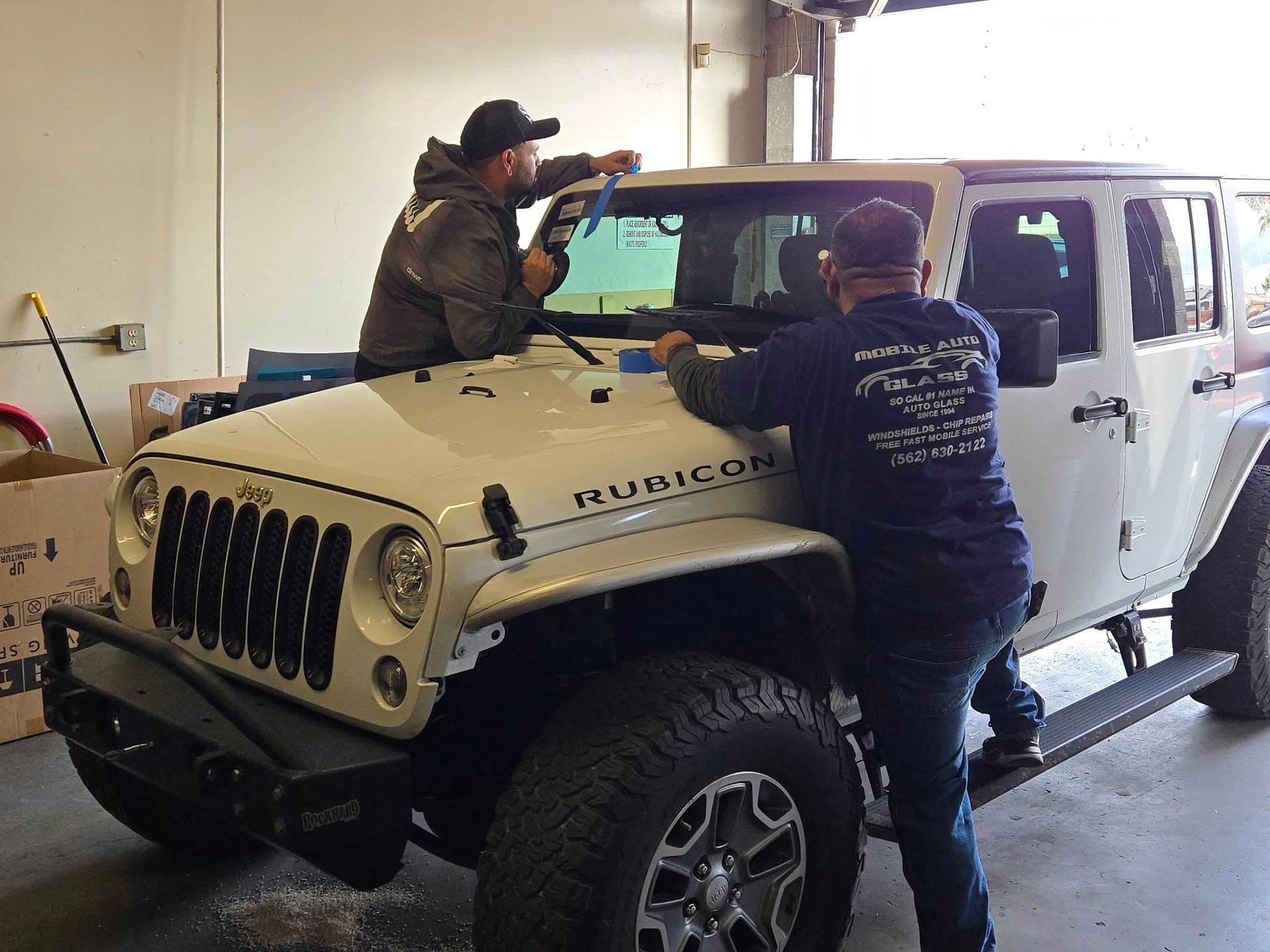 Two men installing a window tint on a white Jeep Rubicon in a garage.