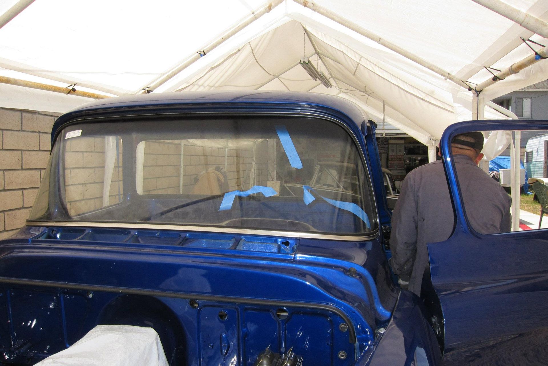 Blue vintage truck cab with windshield being worked on under a tent. A person stands nearby.