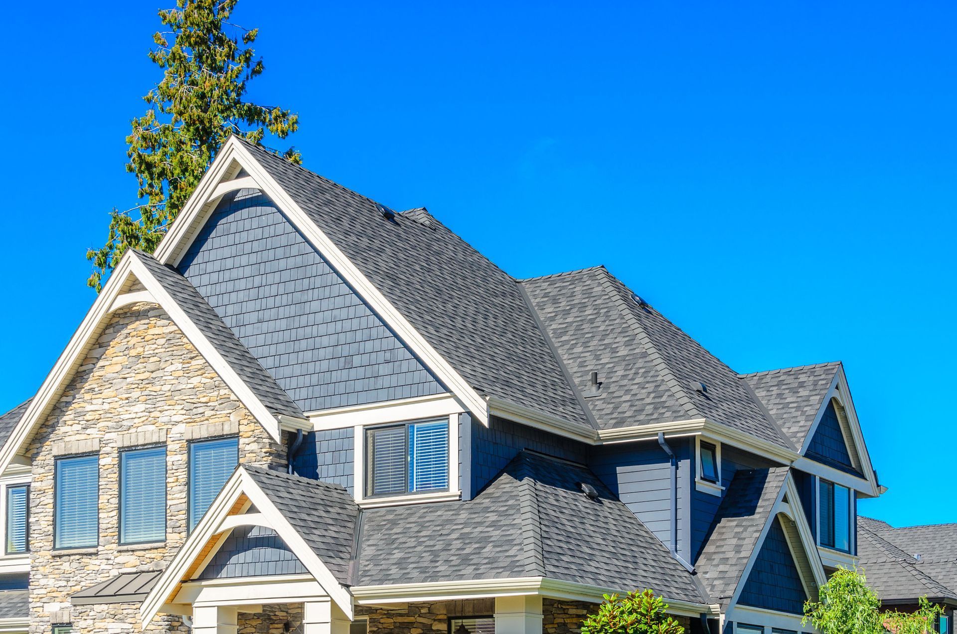 A two-story suburban home with grey shingled roofing, stone siding, and blue accents under a bright clear sky.