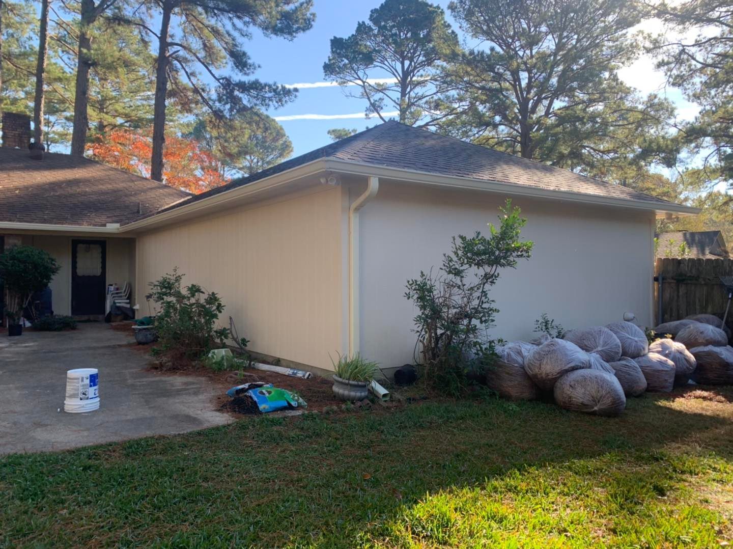 A residential house exterior with light-colored stucco walls, a brown roof, and several bags of soil piled near the corner.