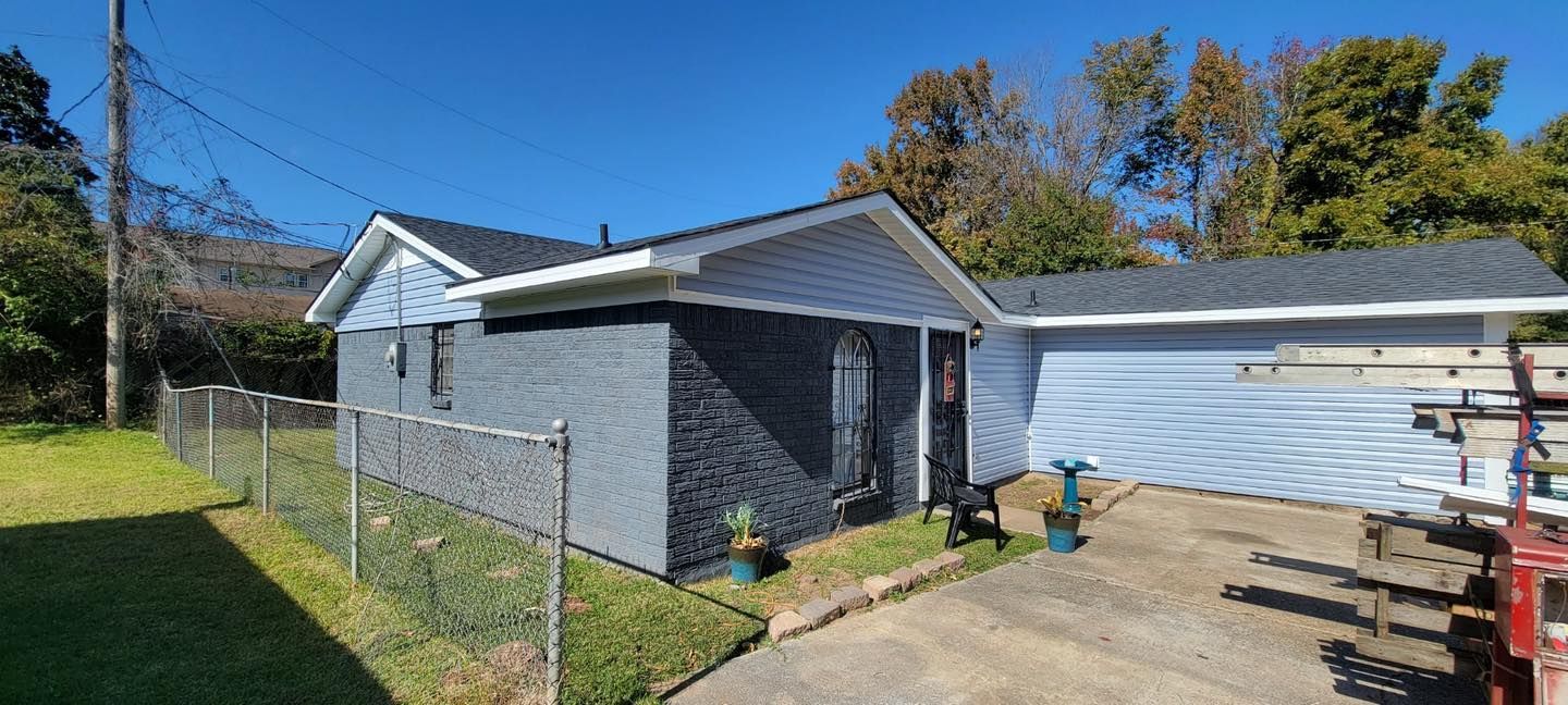 A gray, single-story house with a chain-link fence, a paved driveway, and trees under a clear blue sky.