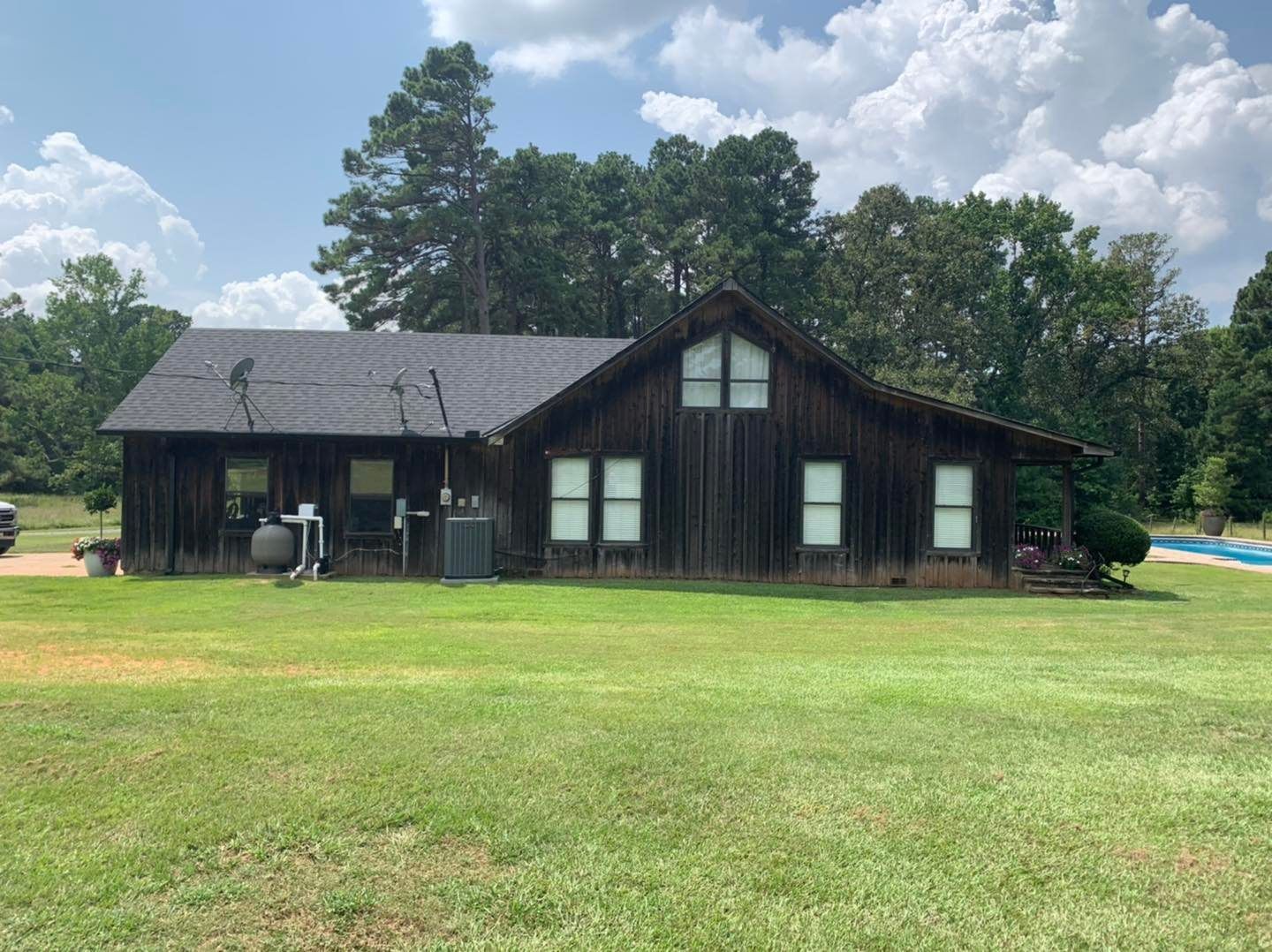 A rustic, dark-wood cabin with a dark shingled roof sits in a large, grassy yard under a cloudy blue sky.