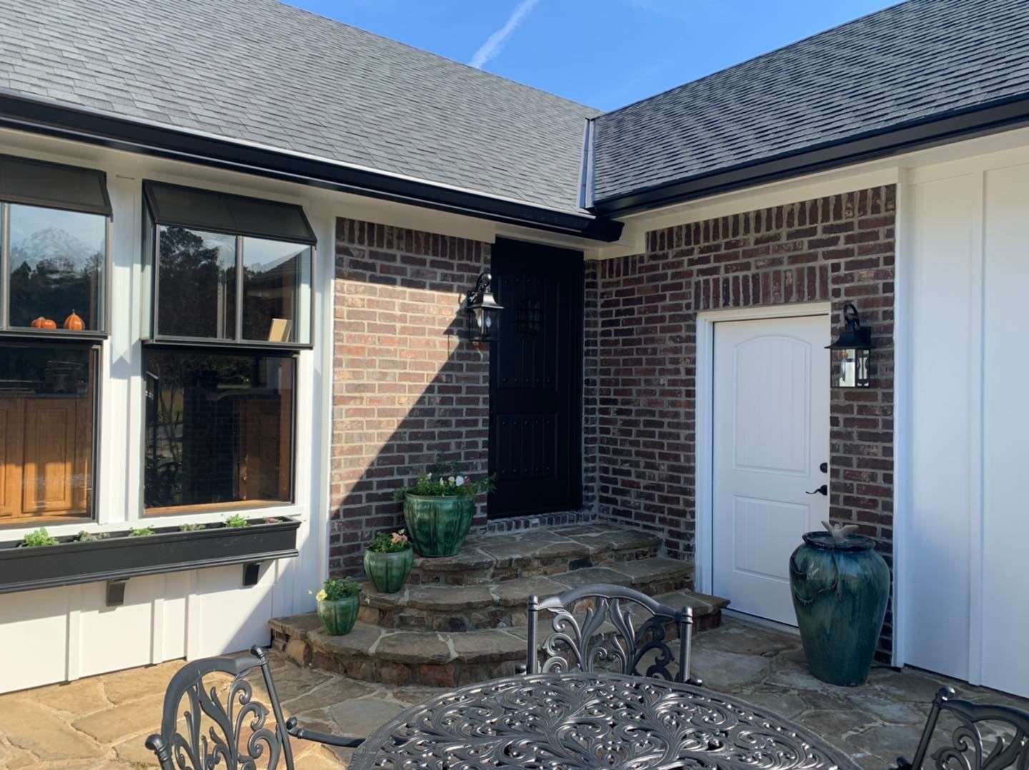 A brick patio area with stone steps leading to a dark door, adjacent to a white door, windows, and decorative planters.