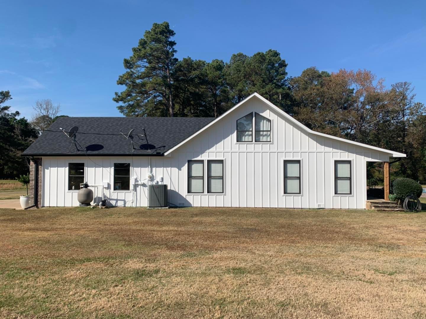 A white, board-and-batten single-story house with a black shingled roof under a clear blue sky, set on a grassy lot.