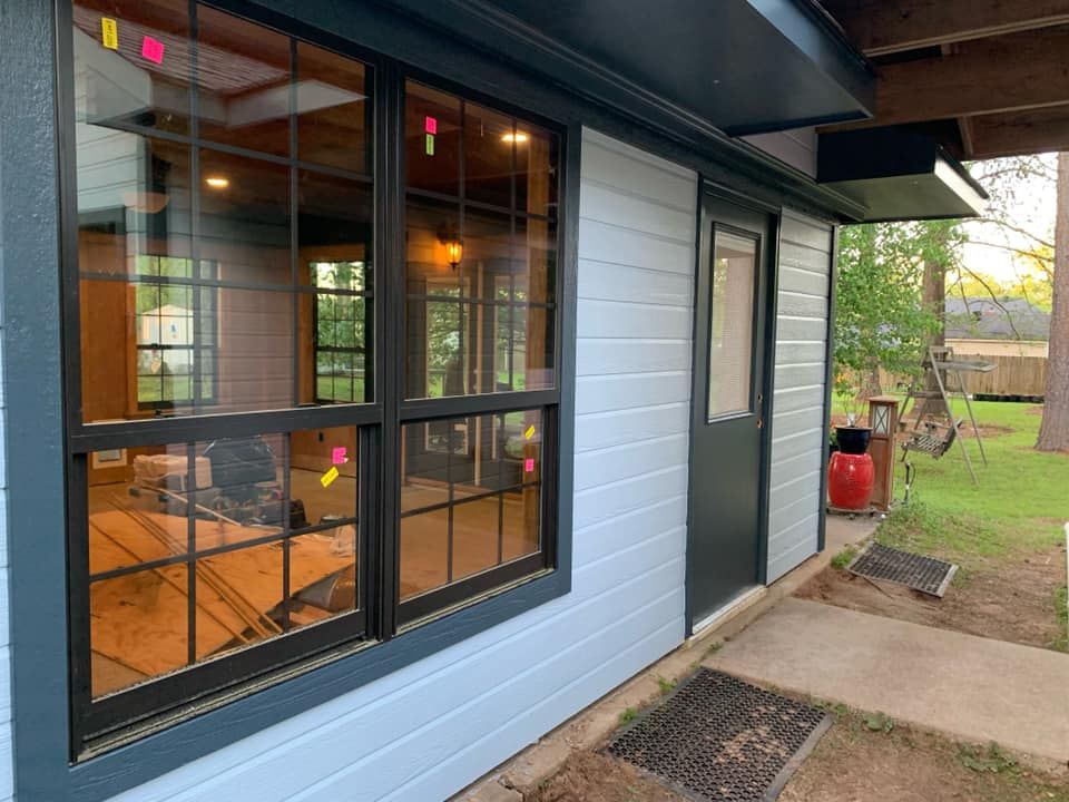 A ground-level view of a light blue house exterior featuring a large, black-framed grid window and a matching entry door.