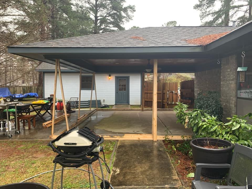A covered patio with a grill in the foreground, wooden support posts, a concrete floor, and a white house in the background.