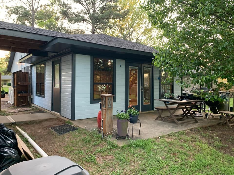 Light blue outdoor space with a wooden dining set on a patio, adjacent to a house with large windows and glass doors.