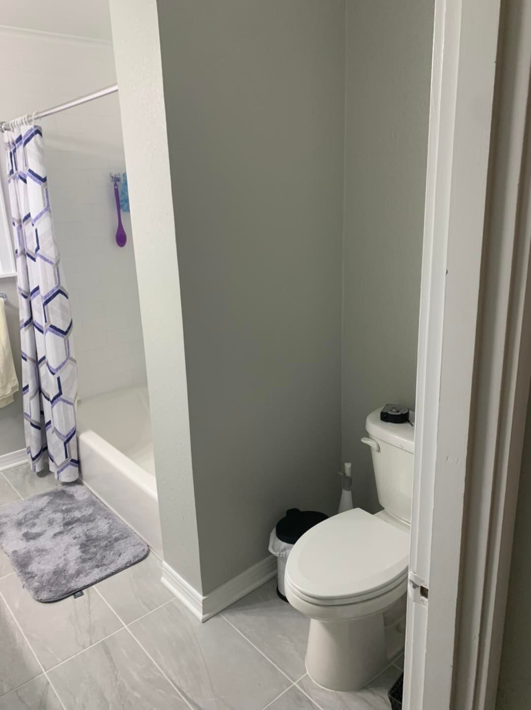 A bathroom featuring a gray wall, a white toilet with a small black trash can, and a shower with a blue-patterned curtain.