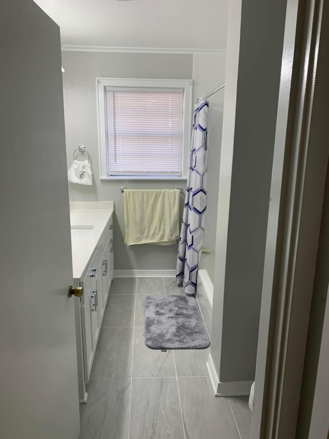 A bright bathroom with light grey walls, white cabinets, a window with blinds, a towel, and a patterned shower curtain.