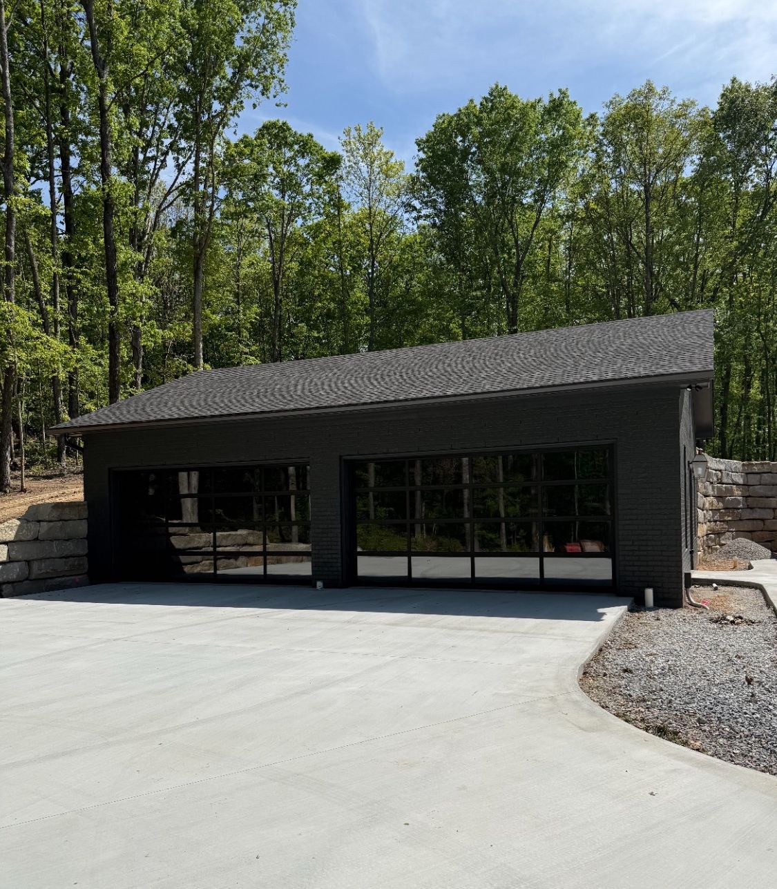 Modern black garage or outbuilding with large glass doors beside a wooded driveway
