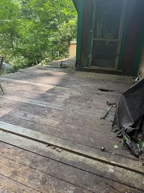 Weathered wooden deck with damaged boards. A screened door is visible in the background, surrounded by green foliage.