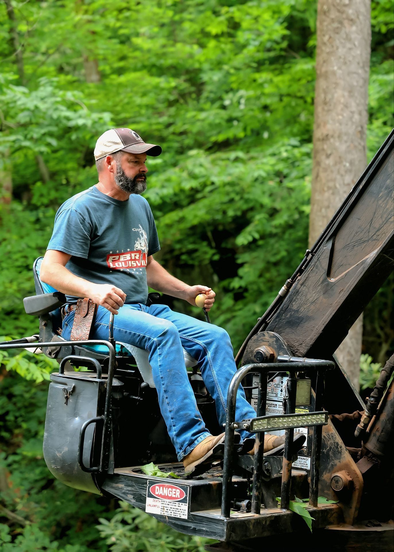 Man in a bucket lift trimming a tall tree in a forest setting; he's wearing a cap, jeans, and a t-shirt.