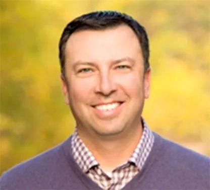 Smiling man in a purple sweater over a checkered shirt against a blurred yellow-green background