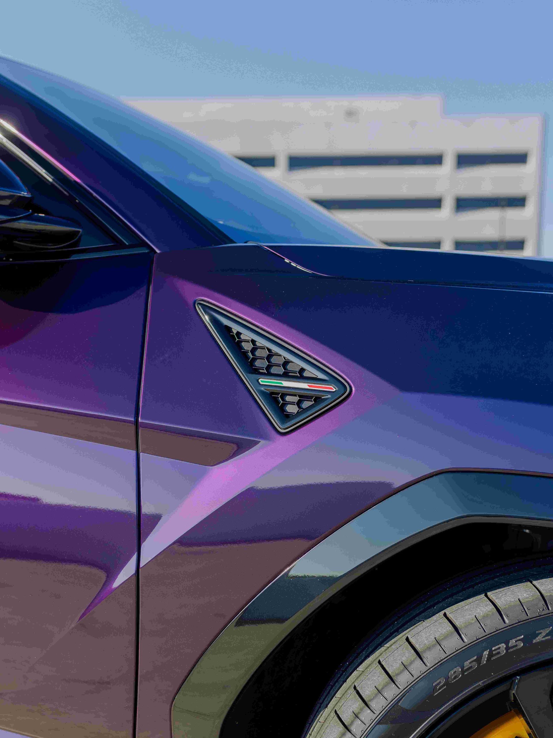 Purple sports car front fender with a triangular badge and visible wheel, parked near a building.