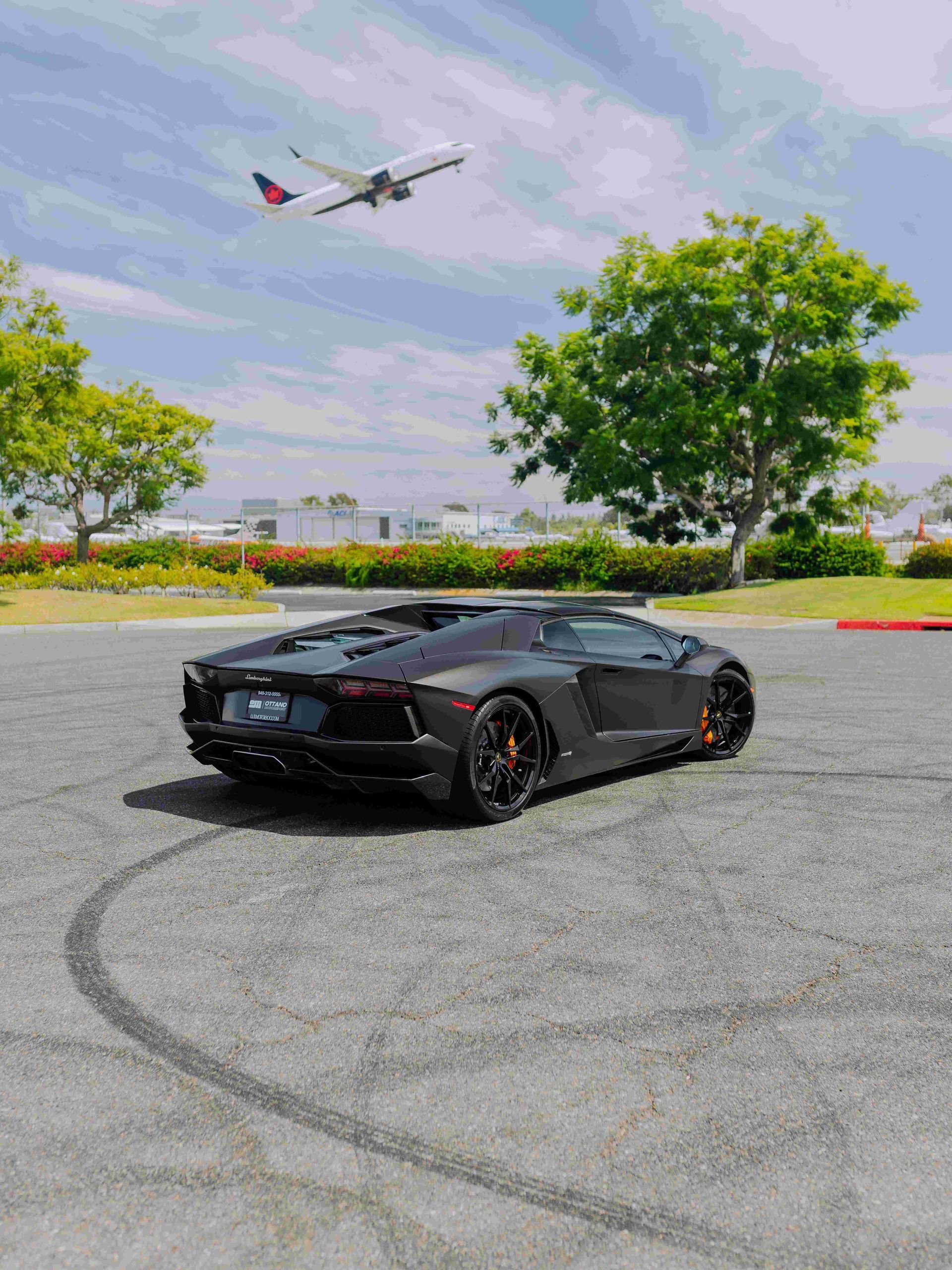 Black sports car in a parking lot with a plane flying overhead and trees in the background