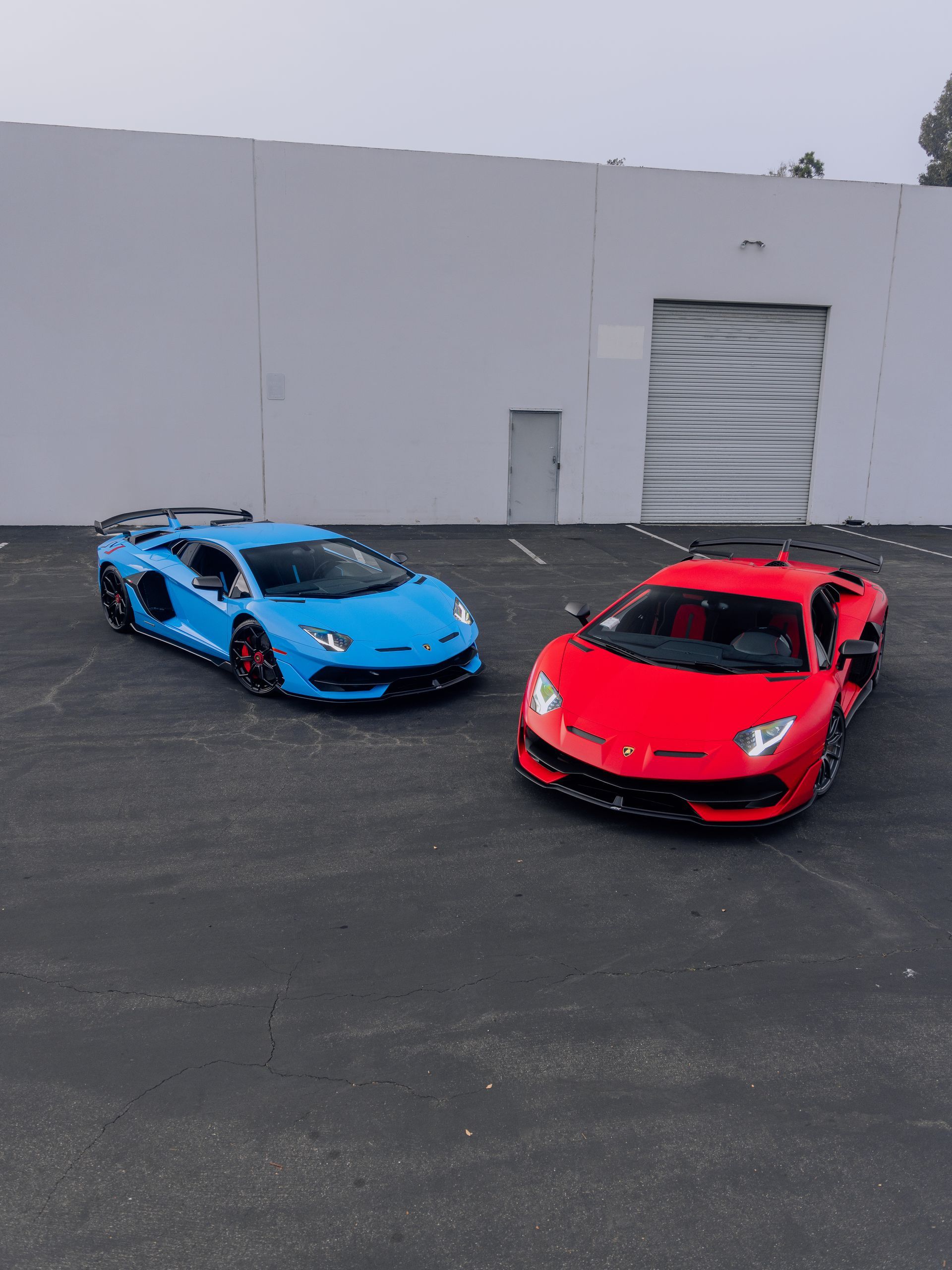 Two sports cars, one blue and one red, parked on asphalt in front of a white building