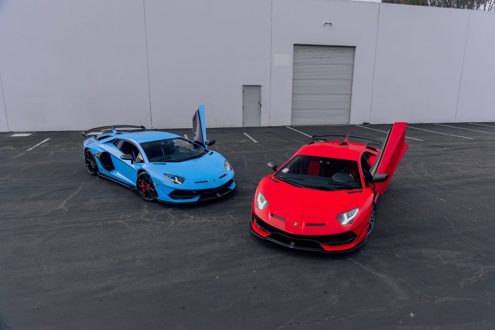 Two Lamborghini sports cars, one blue and one red, parked on pavement with scissor doors open.