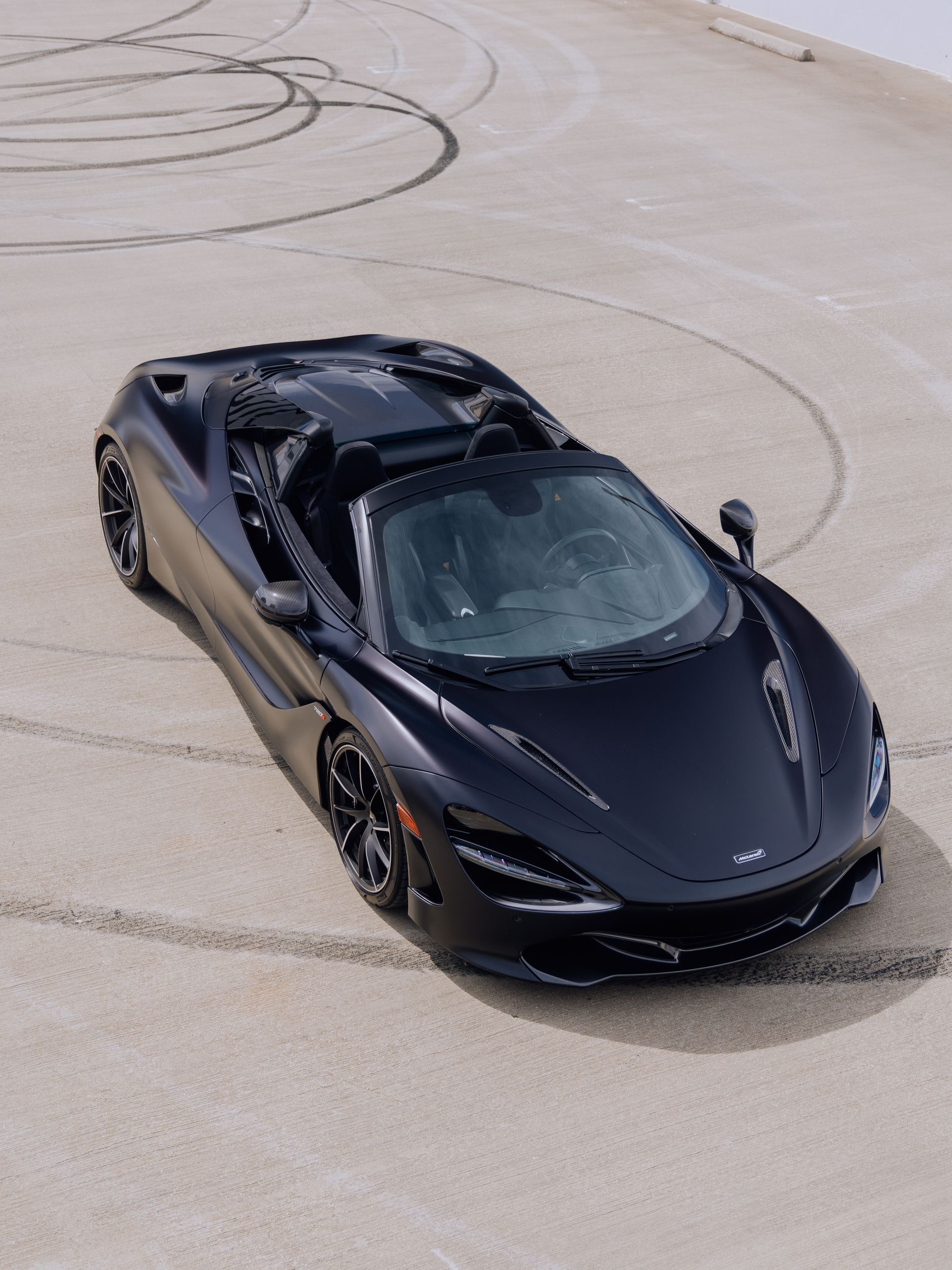 Black sports car driving on a wide paved surface, viewed from above.