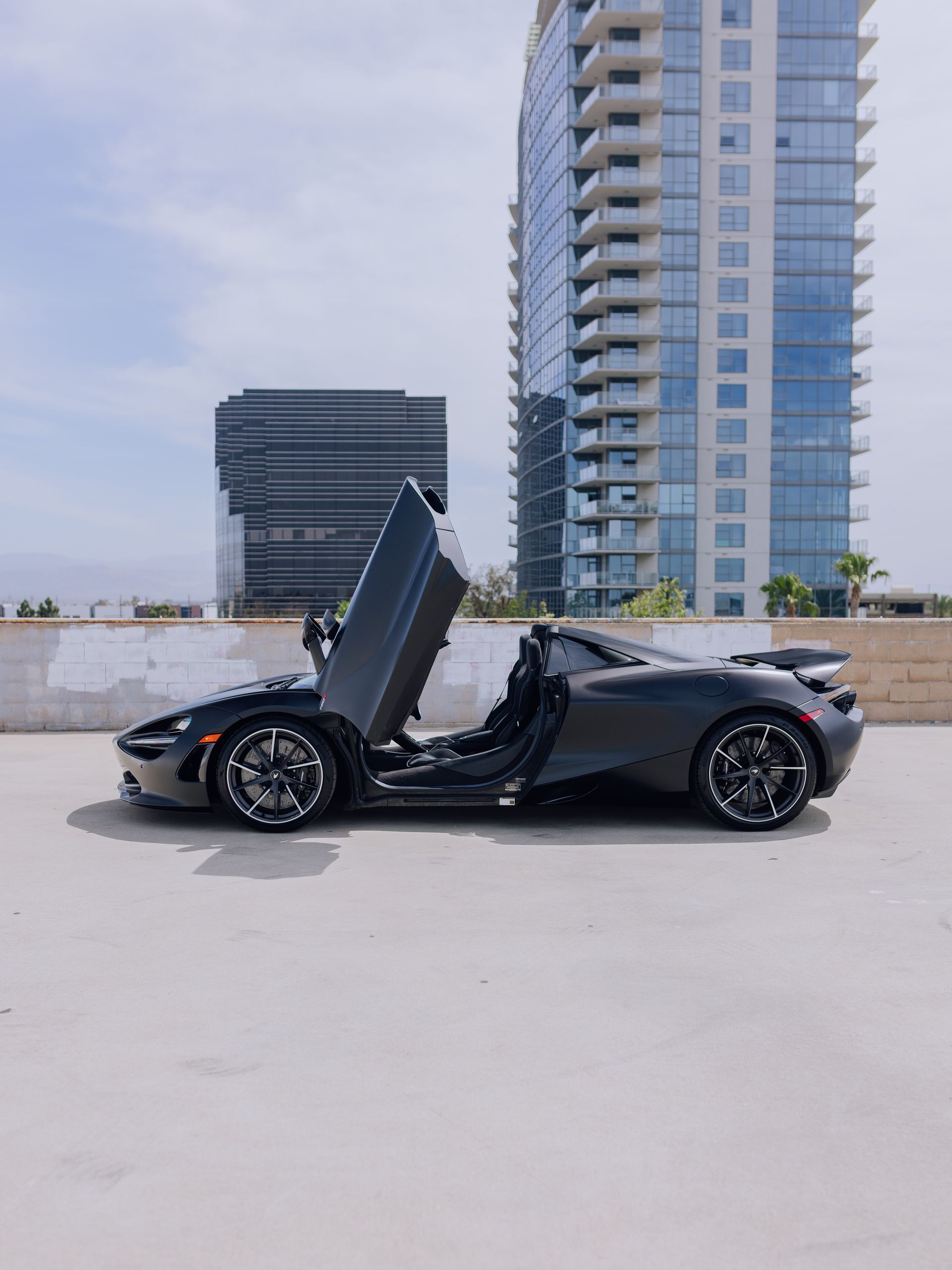 Black sports car with butterfly door open, parked on a rooftop near modern high-rise buildings.