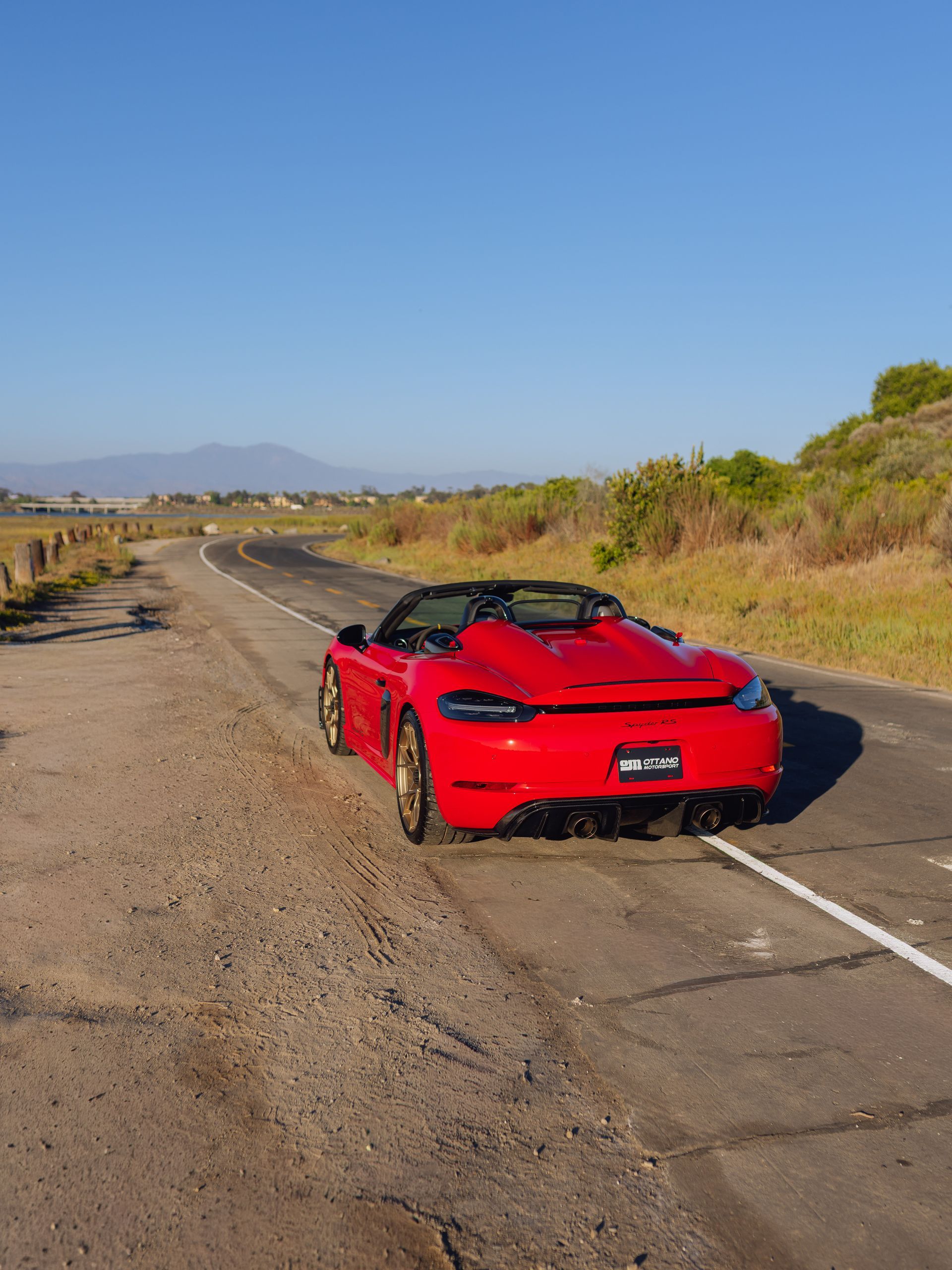Red sports car parked on a roadside under a clear blue sky, with hills in the distance.